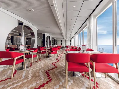 Helles, modernes Restaurant an Bord der Mein Schiff 6. Rote Stühle, Holztische und großer Meerblick durch Panoramafenster. Gemusterter Teppich. Ohne Personen.