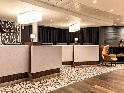 Blick auf die geräumige und modern gestaltete Rezeption an Bord der Mein Schiff 6. Mehrere Tresen, ein Loungesessel und ein stylischer Kamin mit Büchern sind zu sehen.