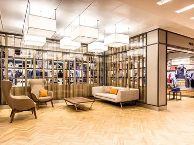 Blick in den eleganten Einkaufsbereich an Bord der Mein Schiff 6 mit beigen Sesseln, Sofa und einem Holztisch. Hinter einer Glaswand sind Kleidung und Taschen ausgestellt.