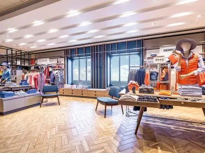 Blick in einen modernen Mode-Shop an Bord der Mein Schiff 6. Mannequins, Kleiderständer und gefaltete Kleidung sind zu sehen. Große Schiffsfenster bieten Meerblick.