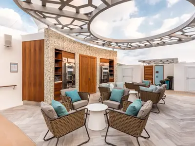 Blick auf das X-Sonnendeck der Mein Schiff 6. Die Lounge-Sessel mit blauen Kissen stehen auf einem Holzboden unter einem offenen Dach. Es gibt zwei Barschränke und Türen. Der Himmel ist bewölkt.