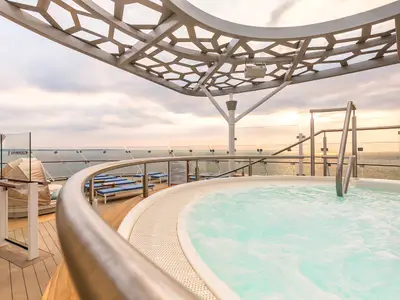 Blick auf einen belebten Whirlpool auf dem X-Sonnendeck der Mein Schiff 6. Dahinter sind das Meer mit Sonnenuntergang und Sonnenliegen unter einer Waben-Überdachung zu sehen.