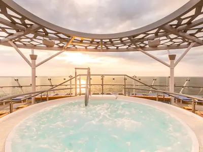 Sprudelnder Whirlpool auf dem X-Sonnendeck der Mein Schiff 6 bei Sonnenuntergang. Das Meer und die warme Lichtstimmung dominieren. Moderne Gitterüberdachung.