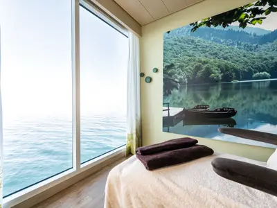 Ein Spa-Behandlungsraum an Bord der Mein Schiff 6 mit Meerblick. Links großes Fenster, rechts Massagetisch mit Handtüchern und Wandbild von einem Bergsee mit Ruderbooten.
