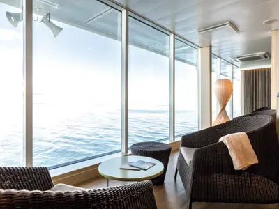 Ein heller Ruheraum an Bord der Mein Schiff 6 mit großen Panoramafenstern, die den Blick auf das weite Meer freigeben. Mehrere bequeme Korbsessel stehen bereit und ein runder Beistelltisch mit Magazinen.