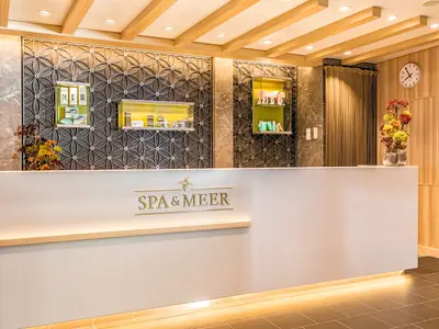 Rezeption des SPA & Meer an Bord der Mein Schiff 6. Weisser Tresen, goldenes Logo. Dahinter Produkte in beleuchteten Vitrinen vor gemusterter Wand. Florale Dekoration und Uhr.