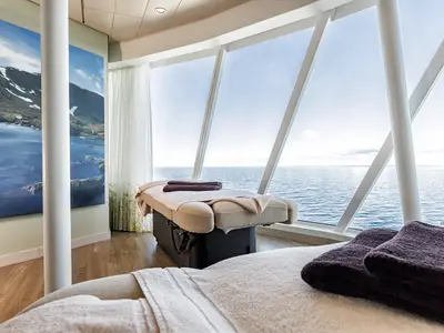 Zwei Massagebetten im hellen SPA & Meer an Bord der Mein Schiff 6. Große Fensterfront mit Panoramablick auf das Meer. Links ein Wandbild mit Berg- und Seenlandschaft.