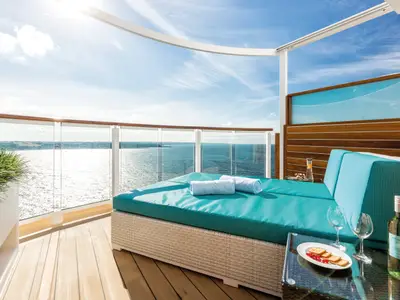 Luxuriöse Entspannungsloge an Bord der Mein Schiff 6: Daybed mit türkisen Kissen, Handtüchern, Wein und Snacks. Im Hintergrund weites Meer und Küste.