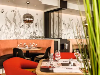 Das GOSCH Sylt Restaurant auf der Mein Schiff 6 zeigt orangefarbene Sitzbänke, Holztische und eine weisse Wand mit maritimem Dekor. Gedeckte Tische mit Gläsern sind zu sehen.