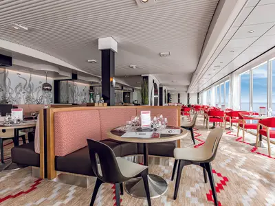 Ein modernes Restaurant, GOSCH Sylt, an Bord der Mein Schiff 6. Der Essbereich bietet rote Stuehle, gepolsterte Sitzbaenke und einen gemusterten Teppich. Grosse Fenster blicken auf den blauen Ozean.