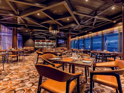 Elegantes Spezialitätenrestaurant an Bord der Mein Schiff 6 bei Abendstimmung. Gedeckte Tische, orange Stühle und großer Meerblick durch Fenster. Moderne Decken- und Lichtgestaltung.