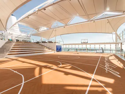 Die überdachte Sportarena an Bord der Mein Schiff 6 zeigt ein Basketballfeld mit Tribüne und Meerblick. Der Boden ist braun mit weißen Linien, überdacht von einem hellen Segeldach.