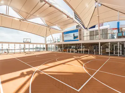 Ein überdachtes Basketballfeld der Arena an Bord der Mein Schiff 6 mit braunem Boden und weißen Linien. Im Hintergrund ist das Meer sichtbar, sowie ein großer Bildschirm mit Informationen und weitere Decksbereiche.