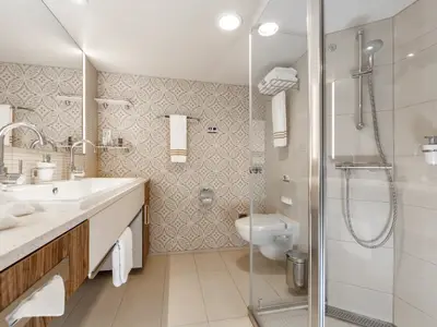 Modernes Badezimmer an Bord der Mein Schiff 6. Es sind ein Doppelwaschbecken, WC, Glasduschkabine und gemusterte Wandfliesen zu sehen. Frische Handtücher liegen bereit.