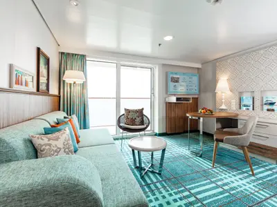 Helle Suite an Bord der Mein Schiff 6. Der Wohnbereich bietet Sofa, Esstisch, Smart-TV und Balkon. Moderne Einrichtung mit Kunst und Obstschale.