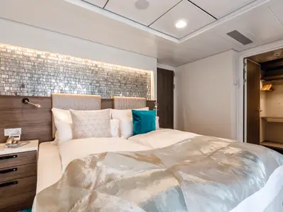 Moderne Suite an Bord der Mein Schiff 6. Grosses Doppelbett, Nachttisch mit Telefon, Mosaikwand über Bett. Offener Kleiderschrank rechts. Helle, komfortable Kabine.