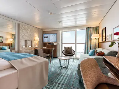 Helle Junior Suite an Bord der Mein Schiff 6. Im Vordergrund ein Bett, dahinter ein Wohnbereich mit Sofa, Schreibtisch und Balkontür. Moderne Einrichtung mit türkisen Akzenten und Meerblick.