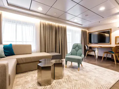 Geräumige Diamant Suite an Bord der Mein Schiff 6 mit großem L-förmigem Sofa, türkisfarbenem Sessel, zwei Metall-Beistelltischen und einem Arbeitsplatz mit TV und zwei Stühlen. Heller, moderner Stil mit Fenstern und Vorhängen.