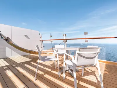 Ein heller Suite-Balkon auf der Mein Schiff 6 mit Holzboden, weißer Sitzgruppe, Hängematte und weitem Blick auf das blaue Meer unter klarem Himmel.