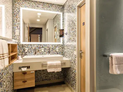 Das moderne Bad einer Himmel & Meer Suite auf der Mein Schiff 6. Es zeigt ein Waschbecken, einen beleuchteten Spiegel und Mosaikfliesen.