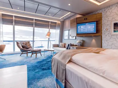 Eine helle Himmel & Meer Suite an Bord der Mein Schiff 6 mit Panoramablick auf das Meer. Zu sehen sind Bett, Sofa, Sessel und TV.