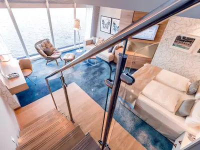 Blick in die zweistöckige Himmel & Meer Suite an Bord der Mein Schiff 6. Zu sehen sind ein Schlafbereich, ein Wohnbereich mit Meerblick und eine Holztreppe.