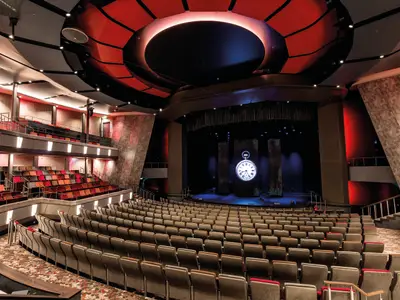 Innenansicht des modernen Theaters an Bord der Mein Schiff 6. Leere Sitzreihen in Grau und Rot. Die Bühne zeigt eine beleuchtete Taschenuhr. Die Decke hat schwarze und rote Elemente.