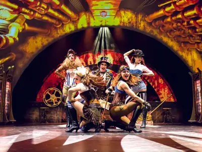 Eine Gruppe von sechs Frauen in detailreichen Steampunk-Kostümen tritt auf der Bühne der Mein Schiff 6 auf. Sie posieren dynamisch unter Scheinwerfern, mit einer Kulisse aus Zahnrädern und Rohren.