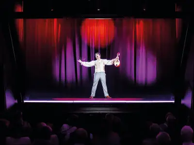 Hologramm-Show im Theater der Mein Schiff 6. Sänger in hellem Hemd, Mandoline, spritzt Wasser auf der Bühne. Rote/lila Vorhänge. Publikum erkennbar.