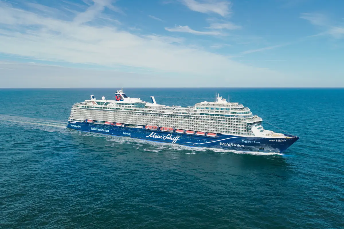 Ein blau-weißes Kreuzfahrtschiff namens Mein Schiff 7 fährt auf blauem Wasser unter einem hellblauen Himmel mit einigen weißen Wolken. Das Schiff ist von links nach rechts in der Mitte des Bildes zu sehen, hinterlässt eine weiße Gischtspur und ist leicht nach rechts gedreht. Die Front des Schiffes ist gut beleuchtet und zeigt die Aufschrift 'Mein Schiff 7'.