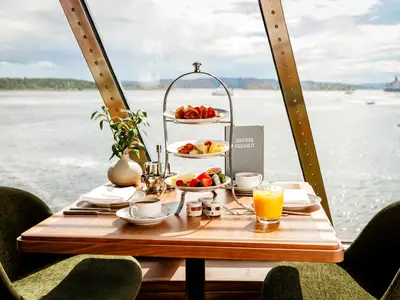Blick aus einem Fenster der Mein Schiff 7 auf das Meer und bewaldete Inseln. Ein Tisch ist mit einem Frühstück für zwei Personen gedeckt, darunter eine Etagere mit Wurst, Käse und Früchten sowie Kaffee und Orangensaft.