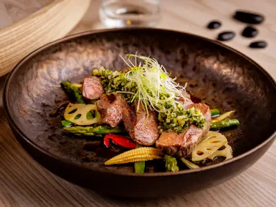 Ein appetitlich angerichteter Teller mit geschnittenem Fleisch, das mit einer Kräutersalsa und feinen Lauchzwiebeln garniert ist, auf einem Bett aus Spargel, Babymais, roten Paprikastreifen und Lotuswurzelscheiben. Das Gericht wird in einer dunkelbraunen, rustikalen Schale serviert und fängt das Essen an Bord der Mein Schiff 7 ein.