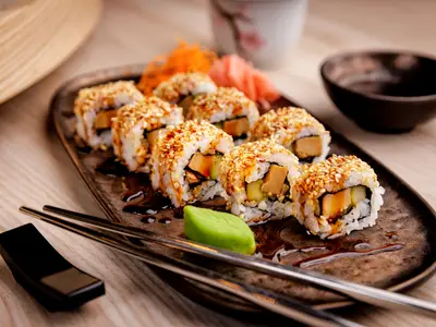 Eine dunkle, ovale Servierplatte mit acht Sushi-Rollen, die mit Sesam bestreut und mit Soße beträufelt sind. Daneben liegen Ingwer, Wasabi und Stäbchen. Im Hintergrund sind weitere Elemente eines gedeckten Tisches auf der Mein Schiff 7 unscharf zu sehen.
