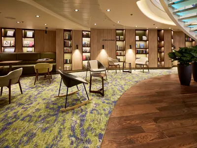 Innenansicht der Leselounge der Mein Schiff 7 mit moderner Einrichtung, Holzregalen voller Bücher, verschiedenen Stühlen, und einem Teppich in Grün- und Blautönen. Rechts ein Bereich mit Holzboden und zwei großen Pflanzen.