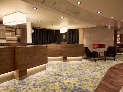 Eine moderne Rezeption auf der Mein Schiff 7 mit warm beleuchteten Holztheken, bequemen Sesseln und einem elektrischen Kamin, die eine einladende Atmosphäre schafft. Der Teppich hat ein unregelmäßiges Muster aus Grün und Lila.