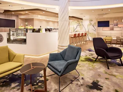 Innenansicht der "Saftwerft" Juice Bar auf der Mein Schiff 7. Es gibt eine weiße Bar mit frischem Obst und Saftpressen. Davor stehen mehrere Sessel in Grün, Blau und Violett sowie Tische. Im Hintergrund sind orange Barhocker und bequeme Bänke zu sehen.