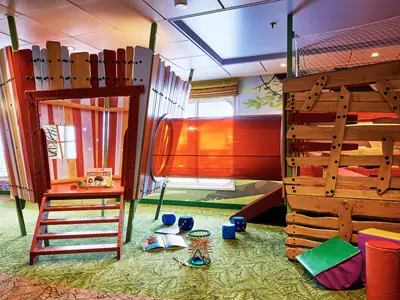Blick in das bunte Spielzimmer im Kids Club der Mein Schiff 7. Es gibt ein Baumhaus mit Treppe und eine orangefarbene Röhrenrutsche. Auf dem Boden liegen blaue Würfel, ein Buch und ein Mikado-Spiel.