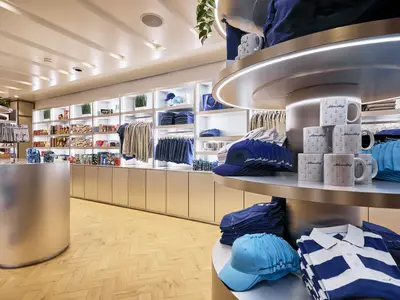 Ein moderner, heller Shop an Bord der Mein Schiff 7 mit Regalen voller Kleidung, Mützen, Tassen mit Anker-Motiv und Snacks. Ein runder Tresen steht im Vordergrund, der Holzboden ist in Fischgrätmuster verlegt.