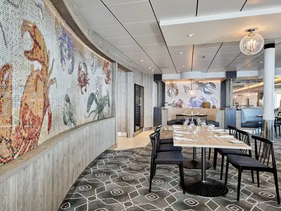 Ein Panoramafoto des Restaurants GANZ SCHÖN DEFTIG an Bord der Mein Schiff 7. Eine lange, geschwungene Wand ist mit maritimen Motiven wie Hummern und Krabben in Mosaikform verziert. Der Raum ist mit Holzakzenten, Teppichboden und Tischen mit schwarzem Stuhl und hellem Holz ausgestattet, fertig eingedeckt mit Gläsern und Besteck.
