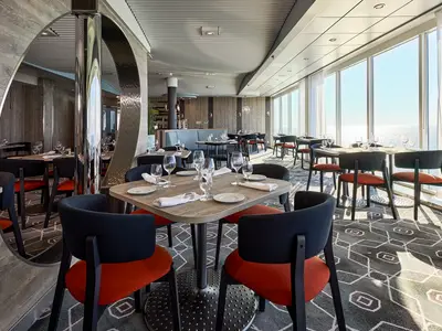 Innenansicht des Restaurants 'Cucina die Stelle' an Bord der Mein Schiff 7. Mehrere Tische mit Stühlen, gedeckt mit Tellern und Gläsern, sind auf einem gemusterten Teppich verteilt. Große Fenster bieten einen weiten Blick auf das glitzernde Meer.