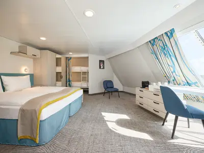 Blick in eine geräumige und helle Kabine der Mein Schiff 7 mit Doppelbett vorne links, Etagenbetten hinten links in einer Nische, einem Arbeitsbereich mit Schreibtisch und Stuhl am Fenster rechts, und einem weiteren blauen Stuhl in der Mitte. Sonnenlicht fällt durch das große Fenster auf den Teppichboden.