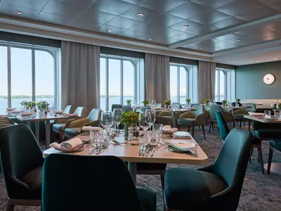 Blick in einen eleganten Speisesaal an Bord der Mein Schiff 7. Mehrere Tische sind mit Besteck, Weinglaesern und frischer Blumendeko gedeckt. Grosse Fenster bieten einen weiten Blick auf das Meer mit bewaldeten Inseln. Die Einrichtung ist in gruenen und beigen Toenen gehalten.