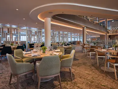 Blick in ein modernes, elegantes Restaurant der Mein Schiff 7. Zahlreiche Tische sind gedeckt mit Besteck, Gläsern und Servietten. Der Raum ist in Grüntönen, Beige und Braun gehalten, mit Säulen, dekorativen Raumteilern und einer oberen Etage mit Blick auf den unteren Bereich. Der Teppich hat ein abstraktes Muster.