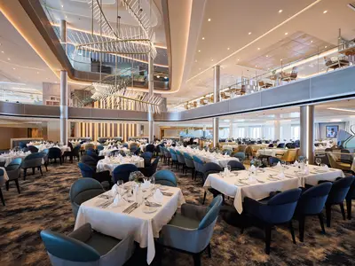 Innenansicht des weitläufigen Hauptrestaurants "Atlantik" auf der Mein Schiff 7, mit zahlreichen gedeckten Tischen, blauen und goldenen Stühlen. Im Hintergrund sind auf einer Empore weitere Essbereiche und ein imposanter Kronleuchter sowie eine Treppe zu sehen.