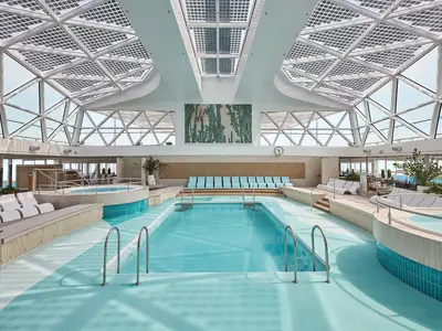 Ein heller, moderner Innenpoolbereich an Bord der Mein Schiff 7 mit einem rechteckigen Pool, zwei Whirlpools und diversen Sitz- und Liegemöglichkeiten. Über dem Bereich befindet sich ein gläsernes Dach mit Solarpanels, das viel Tageslicht hereinlässt. Im Hintergrund ist durch die Fenster das Meer und eine Küstenlinie zu sehen.