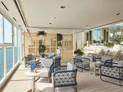 Ein heller Loungebereich an Bord der Mein Schiff 7 mit modernen Sesseln, Holzböden und großen Fenstern mit Blick auf das Meer. Trennwände aus hellem Holz und zahlreiche Grünpflanzen schaffen eine entspannte Atmosphäre. Im Hintergrund ist ein Poolbereich mit Liegen zu sehen.