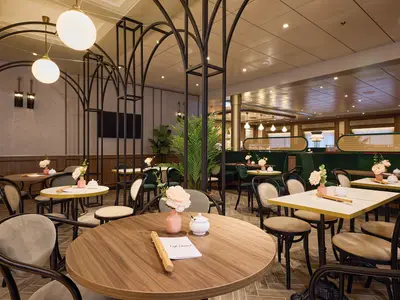 Innenansicht des Café Central an Bord der Mein Schiff 7. Der Raum ist mit eleganten dunklen Holzstühlen, Tischen mit floraler Deko, und grünen Samtsofas eingerichtet. Dekorative Metallbögen strukturieren den Raum, im Hintergrund sind große Fenster zu sehen.
