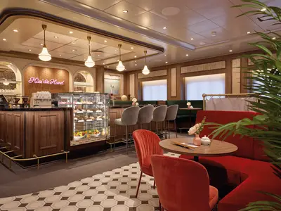Ein elegantes Café an Bord der Mein Schiff 7 mit einer hölzernen Bar, bequemen roten und dunkelgrünen Sitzgelegenheiten, und einer Vitrine voller Gebäck. Die Beleuchtung ist warm und ein Neon-Schild mit der Aufschrift "Küss die Hand" ziert die Wand. Die Einrichtung ist modern-klassisch.