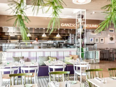 Ein Koch lächelt hinter einer gläsernen Theke in der offenen Küche eines Restaurants auf der Mein Schiff 7. Der Raum ist hell und modern mit Tischen und Stühlen im Vordergrund.