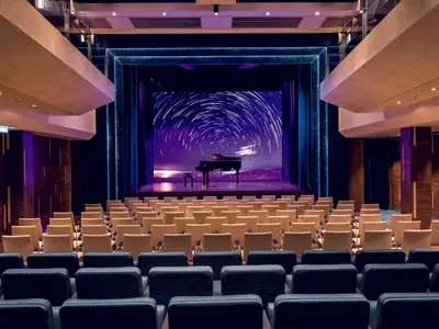 Blick in den leeren Theatersaal der Mein Schiff 7. Reihen von beigen Stühlen stehen vor einer Bühne mit einem schwarzen Flügel. Eine LED-Wand dahinter zeigt eine Sternenspuren-Projektion über einer abendlichen Landschaft. Die Wände sind mit Holzpaneelen verziert.